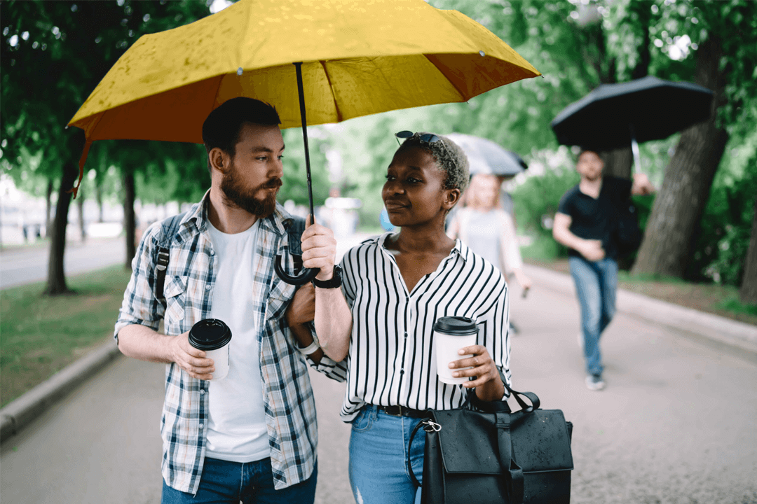 Young multi ethnic casual man and woman walking under light rain in park sharing yellow umbrella and holding cups of coffee at daytime.